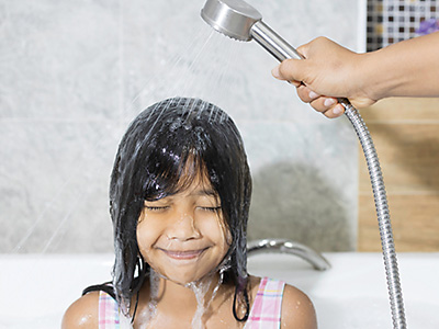 Young girl sitting in bath while mum holds handheld shower head to wash her face and eyes