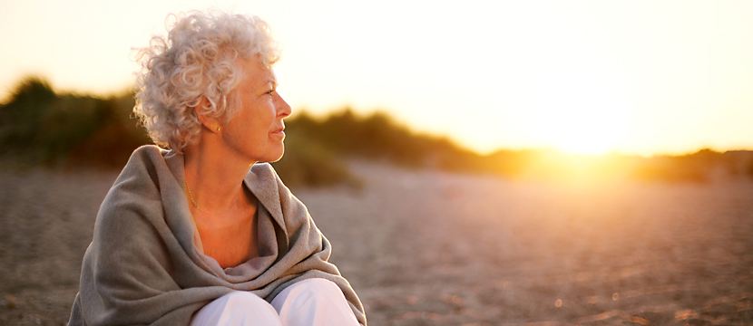 middle-aged woman with curly hair sitting on a beach wearing a shawl with the sun setting behind her