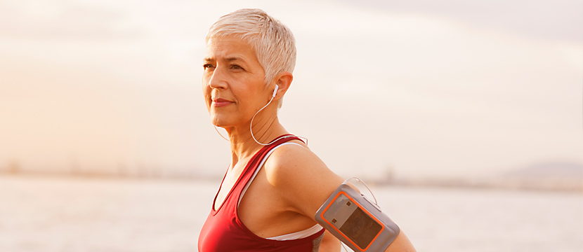 A middle-aged woman with short silver hair in running attire standing on a beach with a phone strapped to her arm and earbuds on.