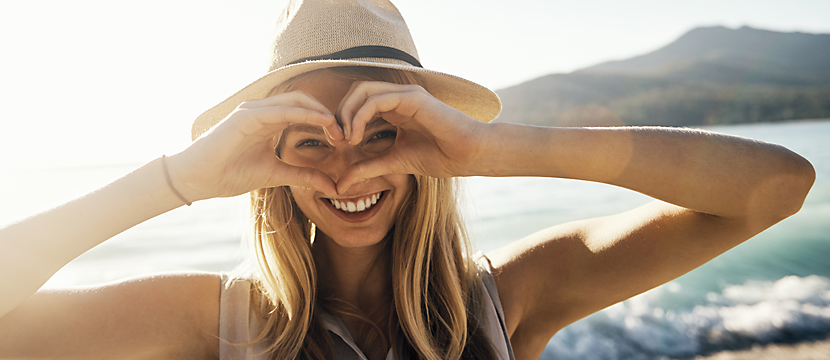 A woman smiling standing on a beach while holding her hands to her face forming a heart shape over her eyes