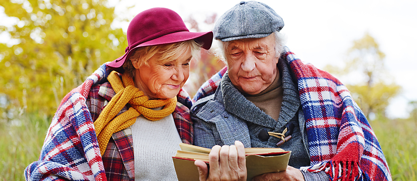 An elderly couple standing together holding a book and dressed for winter with shawls, scarves and hats