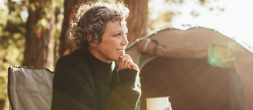 A middle-aged woman smiling and sitting in a camp chair amongst trees and her tent while holding a mug and her chin