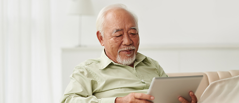 An elderly asian man smiling as he sits while holding an iPad in his hand while reading.