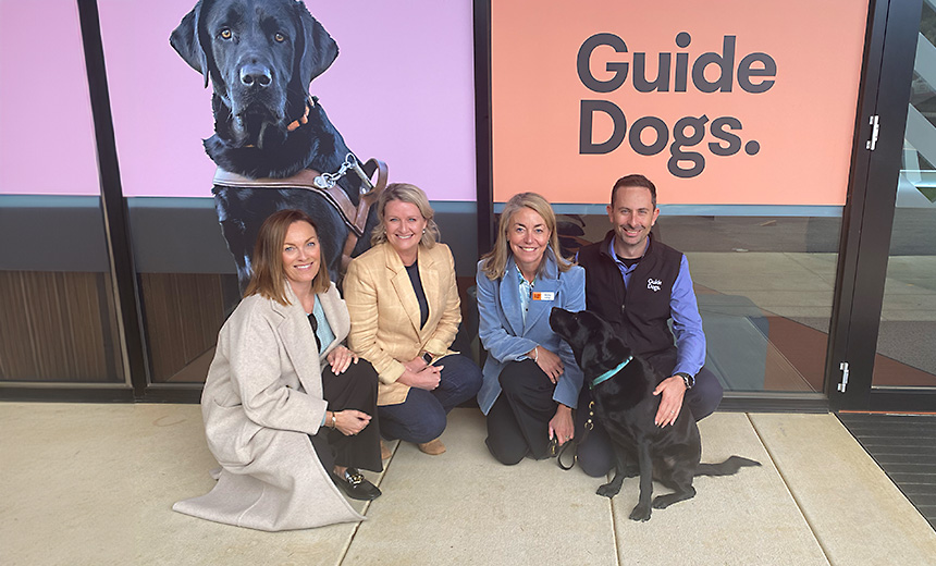 Siobhan Todhunter, Amanda Cranage, Nicky Long and Dr Russell Harrison crouch on the floor in a semicircle with guide dog, Delphi, a black labrador. Delphi is sitting in front of Nicky and Russell, looking up at Nicky. The group are in front of a large sign consisting of a picture of a black labrador on a mauve background next to the Guide Dogs logo (black text on orange background).