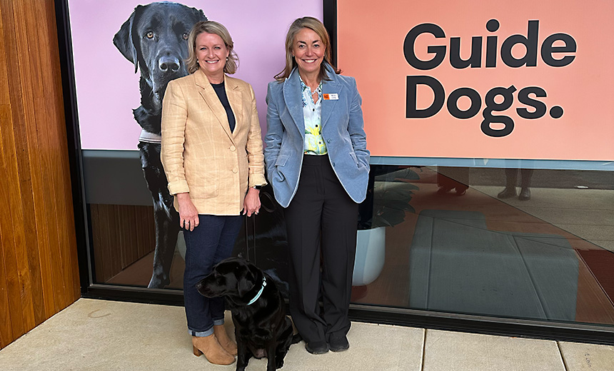 Amanda Cranage and Nicky Long stand with guide dog, Delphi (a black labrador) in front of a large sign depicting a black labrador in a harness on a mauve background, with the Guide Dogs logo on an orange background.