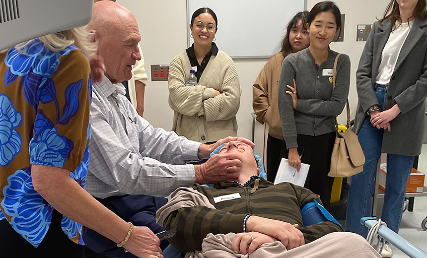 Dr Mark Loane, cataract and refractive surgeon at Vision Eye Institute Brisbane, demonstrates on a pretend patient in the operating theatre at RIverCity Private Hospital, while event attendees observe. Dr Loane has said something amusing, so the attendees and pretend patient are all laughing.