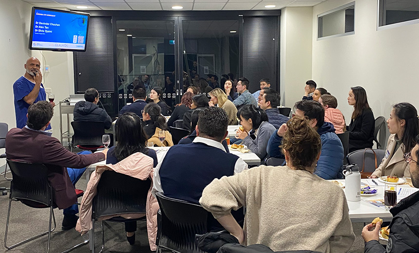 A photo of Dr Devinder Chauhan presenting an opthalmology presentation in a tutorial room. Dr Chauhan is wearing blue scrubs and is holding a glass of red wine while he presents. There is a television screen mounted on the wall behind Dr Chauhan which is displaying a blue and white slide from his presentation. The room is filled with approximately 20–30 young professionals who are sitting around tables. Members of the audience are also drinking glasses of red wine and some are eating small savoury pastries.