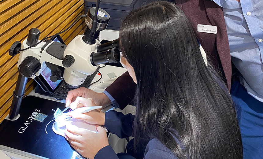 An image of a woman sitting at a desk and looking down a microscope. She has long black hair and is wearing a navy blue jacket. She is holding a white medical device in one hand which she is manipulating under the microscope. There is a man beside her who is holding a pair of tweezers under the same microscope, he appears to be using the tweezer to point something out to her.