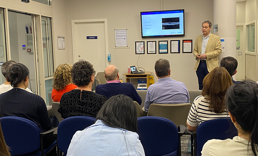 A photo of Dr Eric Mayer presenting an ophthalmology lecture. Dr Mayer is standing in a doctor's office reception area and he is wearing blue chinos, a white shirt and a mustard jacket. He is holding a television remote with both hands. There is a television mounted to the wall behind Dr Mayer. The television is displaying a blue and white slide from his presentation. The room is filled with eleven professionals who are seated in chairs and listening to his presentation.