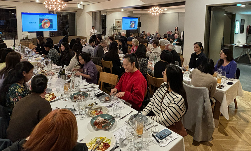 A photo of a large function room with three long tables covered in white tableclothes. There are approximately 50 people seated along these three tables. The tables are set with cutlery and wine glasses and there are half finished meals and wine bottles scattered across the tables. In the background there is a black podium. Dr Christolyn Raj is speaking at this podium. There are two televisions mounted to the wall behind Dr Raj that are displaying a slide titled 'mivision'.