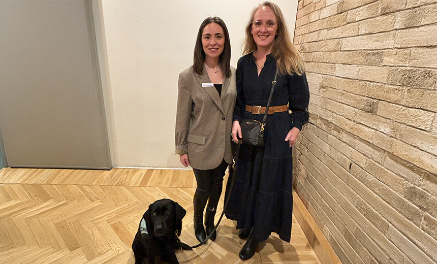 A photo of two women standing next to each other in a room. The women to left is wearing a black top, black jeans, black boots and a beige blazer. The woman to the right is wearing a navy maxi dress with a light brown belt, black boots and a black cross-body bag. The woman in the navy dress is holding a dog leash. Behind the women is a black golden retriever puppy wearing a light blue dog sweater. Both women are smiling.