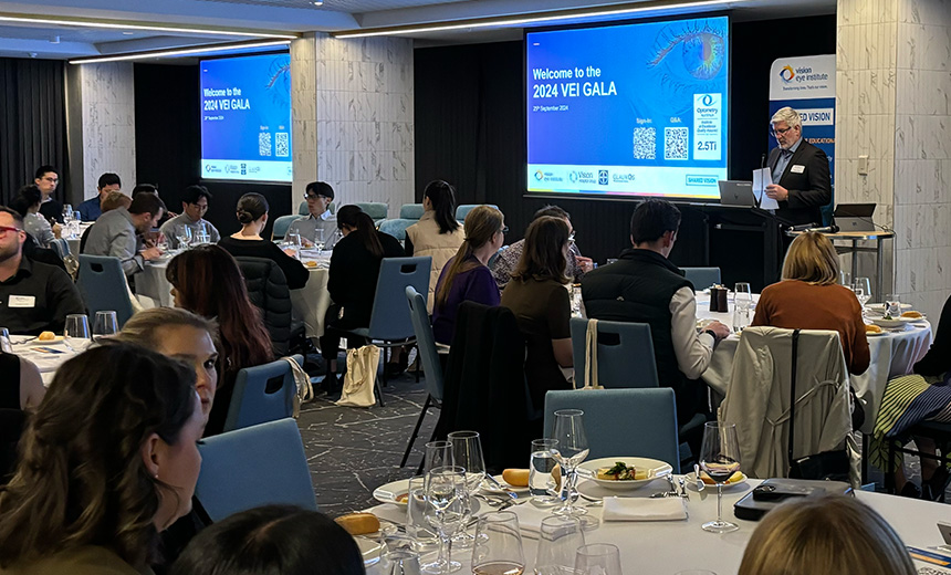 A photo of a group of people seated at round tables in a large hall. There is a man who is wearing a black suit standing at a black podium at the front of the hall. He is holding a white piece of A4 paper and appears to be giving a speech. There is a drop down screen on the wall behind the man which is displaying a blue slide titled 'Welcome to the 2024 VEI GALA'.