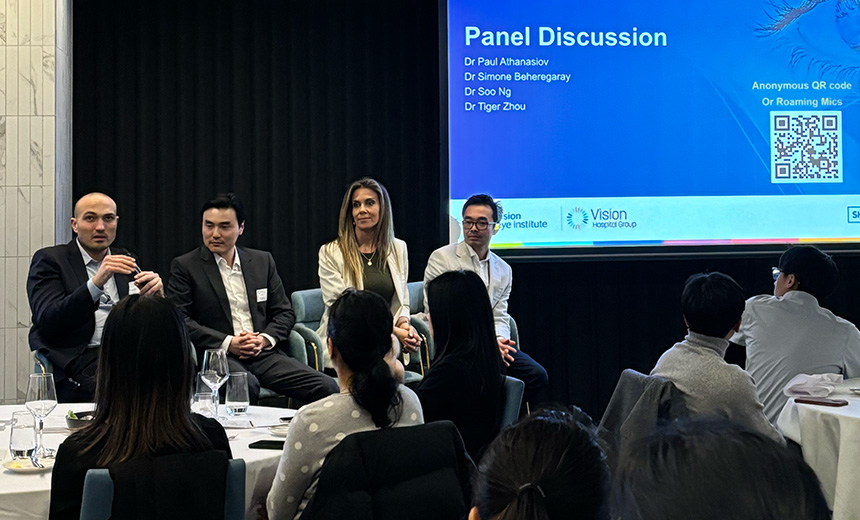 A photo of Dr Paul Athanasiov, Dr Simone Beheregaray, Dr Soo Khai Ng and Dr Tiger Zhou seated in blue velvet chairs at the front of a hall. They are all dressed professionally in suits. Dr Athanasiov is holding a microphone and speaking. There is a drop down screen behind the doctors displaying a blue and white slide titled 'Panel Discussion'. The hall is full of round clothed tables with people seated round them.