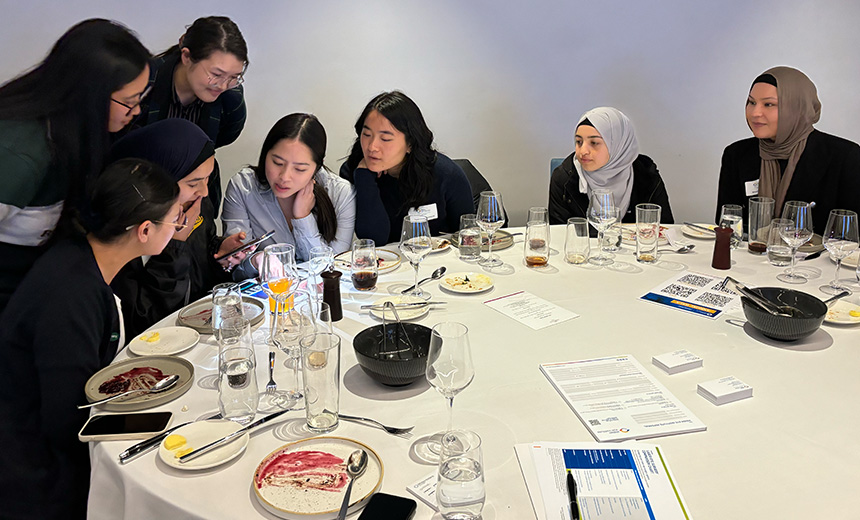 A photo of eight women seated at a round function table. There are glasses and finished meals on the table. The woman are all clustered around one woman's phone and talking.