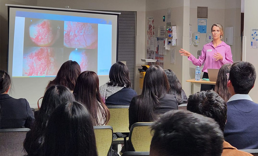 A photo of Dr Simone Beheregaray standing at a podium in an ophthalmology clinic reception area. She is wearing a pink blouse and black pants. There is a screen against the wall besides Dr Beheregaray which is displaying a slide filled with ophthalmology images. There are people seated in front of Dr Beheregaray listening to her presentation.