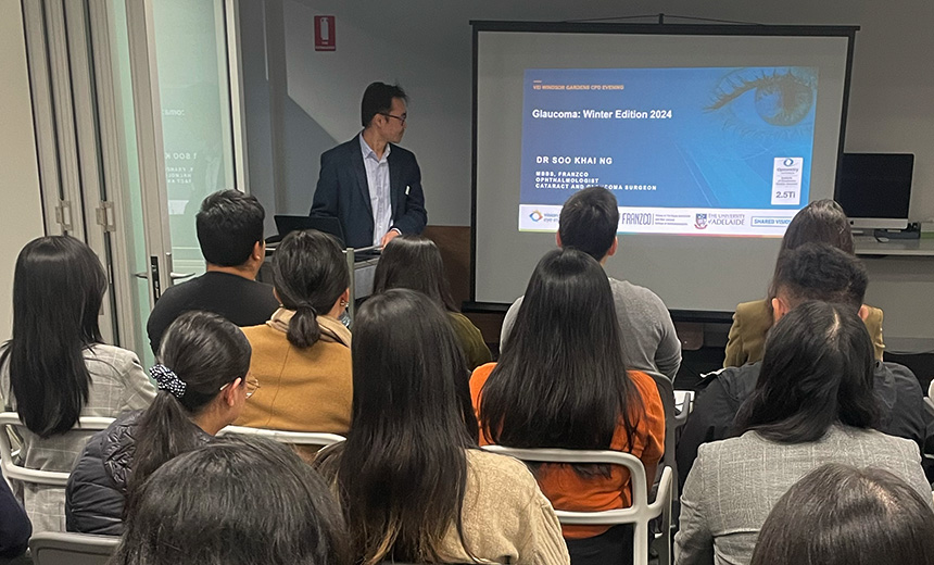 A photo of Dr Soo Khai Ng presenting an ophthalmology lecture in a tutorial room. Dr Ng is standing in front of a metal trolley which a black laptop is placed on. He is turning towards the wall behind him where a large screen is placed. The screen is displaying a blue and white slide titled 'Glaucoma: Winter Edition 2024'. There are seats organised in rows in front of Dr Ng filled with people attending his presentation.