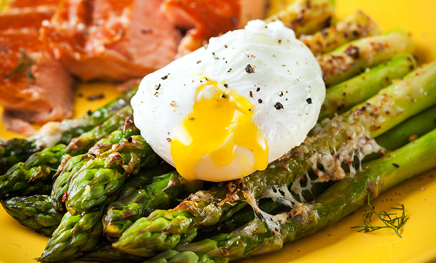 Half a dozen asparagus spears topped with a poached egg sit on a bright yellow plate. The egg has been pierced so that the bright yellow yolk is visible, echoing the colour of the plate. In the background, browny-pink pieces of poached salmon can be seen.