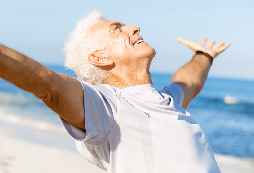 A man with white hair smiling as he opens his arms wide while standing in front of the sea