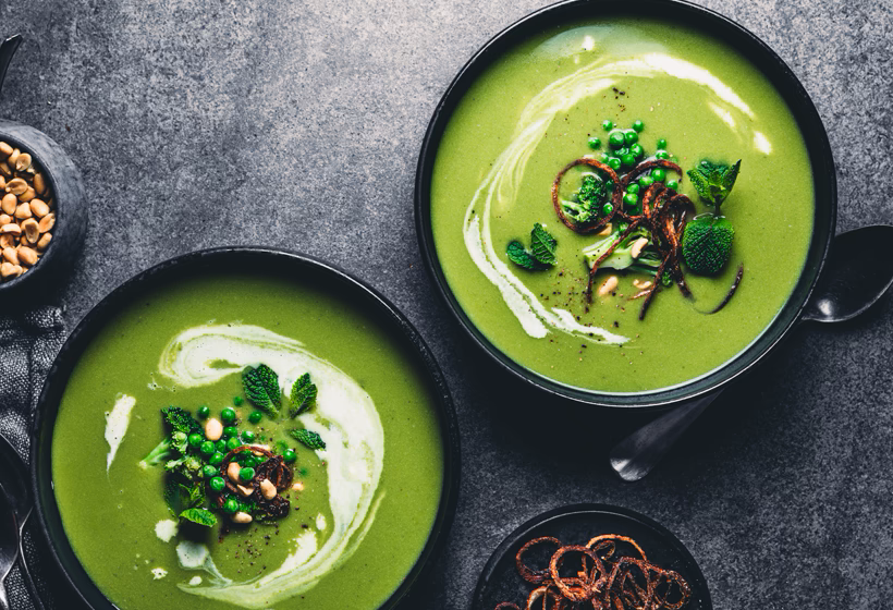 Two bowls containing green broccoli soup, garnished with peas and mint