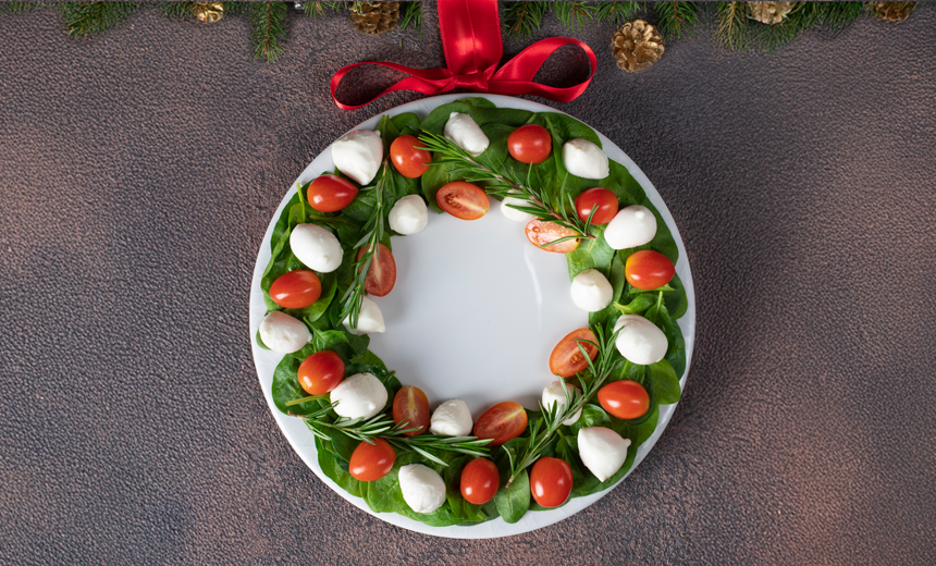 Basil leaves, small bocconcini balls and cherry red tomatoes arranges on a white circular platter in the shape of a wreath. A red fabric bow is positioned at the top end of the plate, and the plate is resting on a table.