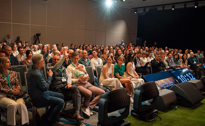 Image of the audience at AUSCRS looking out from the centre left of the stage. It's an indoor conference room with a brown, panelled wall at the back, you can see the tele-prompter and time screens, the time reads 10:12
