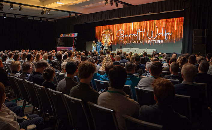 AUSCRS conference room shot, taken from the back right of the room at the start of the Barret Wolfe gold medal lecture – you can see the Barret Wolfe title screen at the back of the stage. The background is gold/bronze streamers overlaid with a gold medallion to the left, the Barret Wolfe in white, cursive writing and 'Gold medal lecture' in all caps underneath
