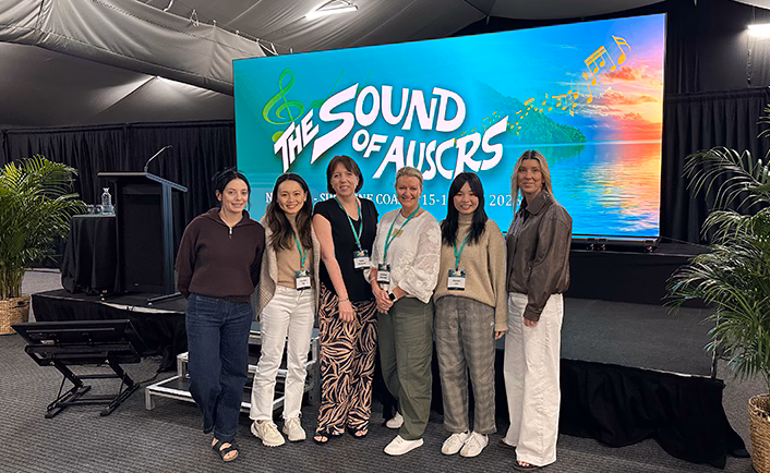 Group shot of Vision Eye Institute (VEI) CEO Amanda Cranage (third from the right) with other VEI staff (5 others) at AUSCRS in front of the 'The Sound of AUSCRS' LED screen (visual like the 'The Sound of Music' movie title card but with a turquoise background and tropical island instead of mountains