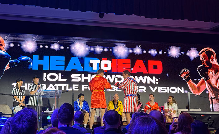 Shot of the main stage for AUSCRS head-to-head session, an 'IOL showdown'. The presenting doctors are dressed in theme as boxers wearing 'costume robes' and the moderators dressed in black and white as referees. Two doctors are standing and 'facing off' the doctor on the left is wearing a red satin boxer robe with yellow trim and belt, the doctor on the right is wearing a red and white vertical striped satin boxer robe with cobalt blue trim and belt with white stars, remaining speakers/contestants are sitting in chairs at the back of the stage. The title screen reads 'Head to Head, IOL showdown: the battle for better vision' with stock images of boxers book-ending the screen.