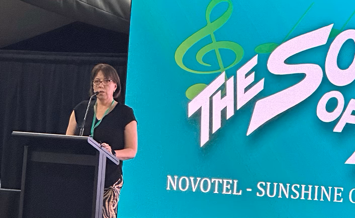 Close up shot of a Vision Eye Institute key speaker at the 'Sound of AUSCRS' staff support program. The key speaker is a woman with brown hair in a bob and glasses, wearing a black t-shirt and standing behind a lectern. You can see the bottom left of the 'The Sound of AUSCRS' title screen which is set up to look like the 'Sound of Music' movie title card