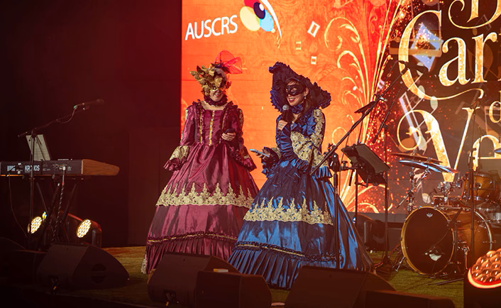 Close up shot of the AUSCRS stage which has been set up for the Gala with band on stage, you can see a drum kit to the centre back and a keyboard to the left. Two women on stage are dressed in full Venetian masquerade gowns with black lace face masks. The lady on the left (facing out to the audience) is in a burgundy satin gown with gold trim, a gold face mask and large gold fascinator, the women on the right is in a royal blue satin gown with gold trim, a black lace mask and large blue hat black lace trim. You can see part of the themed screen which is orange and black with AUSCRS logo and part of the event name 'Biennale Carnevale di Venezia'