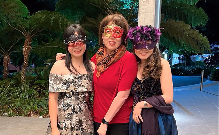 Group shot of three Vision Eye Institute (VEI) staff dressed up in gowns and masked for the Venetian Carvivale Gala dinner. Woman on the left wears an off-the-shoulder black and white floral gown and black and red lace eye mask, the woman in the centre is wearing a red short-sleeved top with black dress pants, an orange and black neck scarf and red and gold face mask, the woman on the right is wearing a blue and purple gown with a black embroidered bodice with blue, purple and gold thread, and blue velvet skirt, purple shawl draped across her arm and a purple face mask with black and purple feathers at the top. The women are outside the venue.