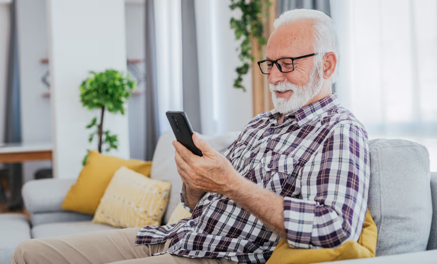 An older man with white hair and a short white beard, wearing glasses and a plaid shirt, smiles while looking at a smartphone. He is seated comfortably on a grey sofa in a bright, modern living room.