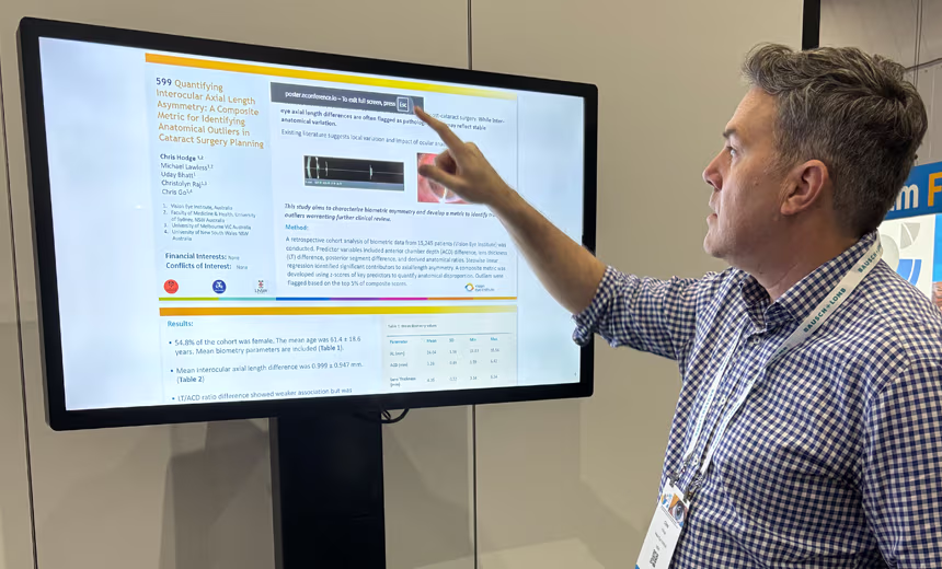 Close up image of Vision Eye Institute’s research coordinator Dr Chris Hodge (PhD) presenting research at RANZO. A slide deck presentation is displayed on a TV in the background, Dr Hodge in a navy blue, gingham, collared-shirt checked shirt faces towards the screen (away fro the audience) and points at a key point at the top of the visible slide on the TV.