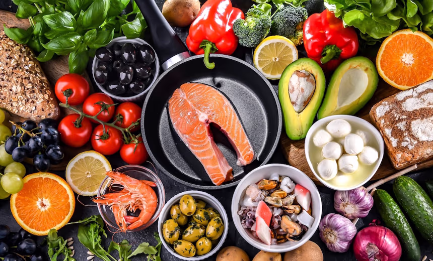 An overhead flat-lay of a variety of nutrient-rich foods associated with eye health, including a raw salmon steak in a frying pan, prawns, mussels, olives, avocado, tomatoes, red capsicum, broccoli, citrus fruit, grapes, leafy greens, garlic, mozzarella, and wholegrain bread, arranged on a dark surface.