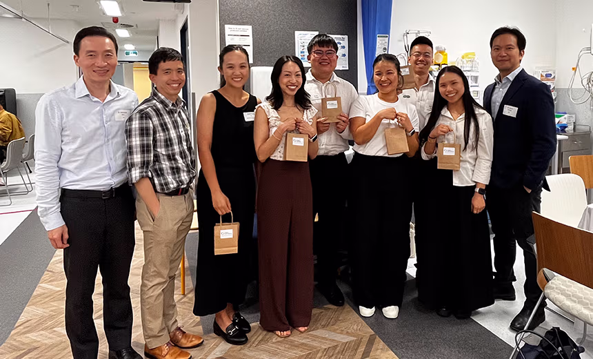 A group of nine people smiling and posing together in a clinical or medical setting, several holding small branded kraft paper gift bags. The group includes a mix of ophthalmologists, including Dr Michael Wei, Dr Christopher Go and Dr Jason Cheng, along with referring optometrists.