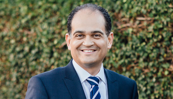 Dr Alex Ioannidis dressed in a suit and tie outside in front of a ivy covered wall