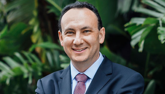 Dr Lewis Levitz dressed in a suit and tie standing in front of a wall of palm trees