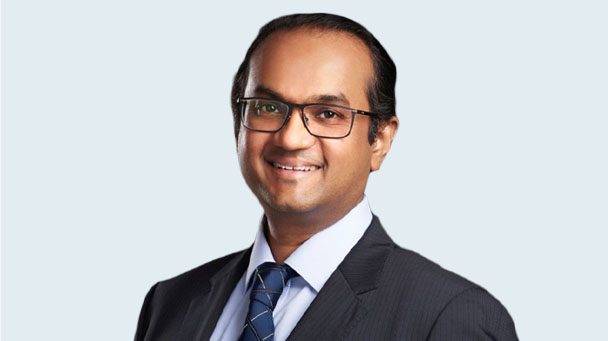 Headshot of ophthalmologist Dr Deveraj Supramaniam. He wears a dark charcoal suit, dark blue tie, white-collared shirt and glasses. He is standing in front of a pale blue wall.