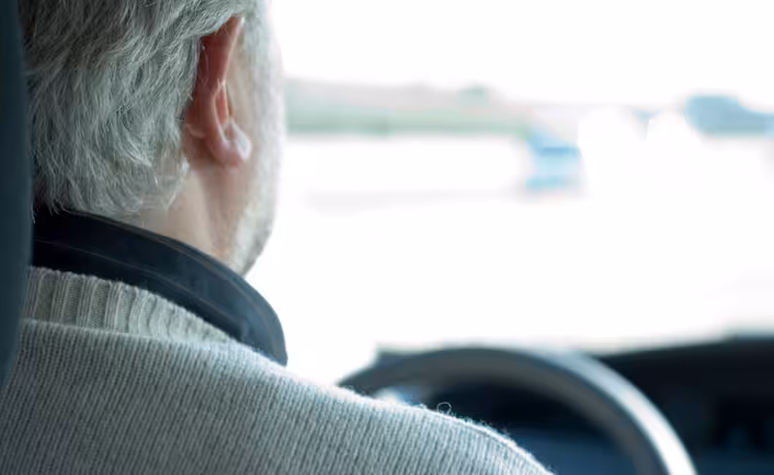 Back view of a senior man with gray hair driving a car, hands on the steering wheel, looking out at a blurred road ahead.