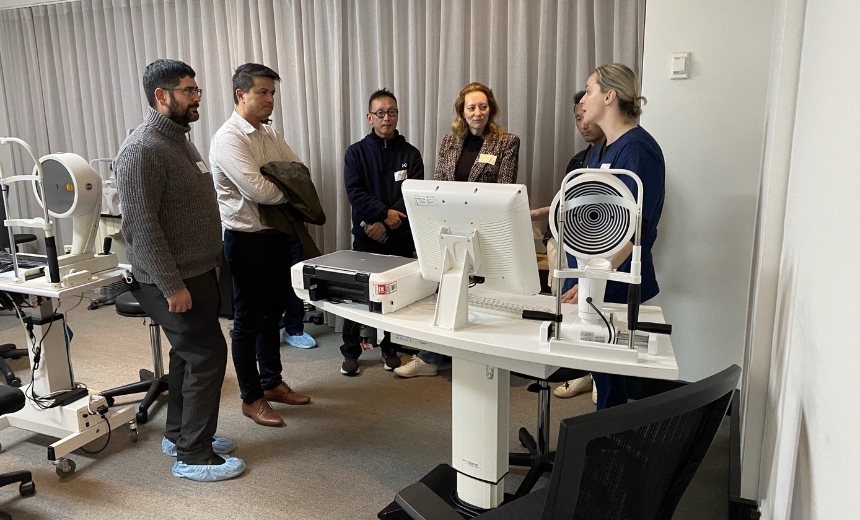 Six people attending a CPD education event at the Vision Eye Institute Melbourne clinic. They are all standing around a desk. Computer equipment is placed on the desk, and the attendees are in discussion with one another.