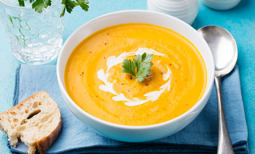 Bowl of pumpkin soup, garnished with cream and a sprig of parsley, served with slice of crusty bread on blue table setting