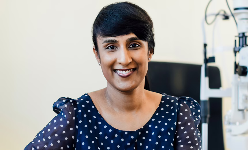 Dr Rushmia Karim (woman) in consulting suite, next to a microscope, wearing a navy blue dress with white polka dots; hair in a ponytail