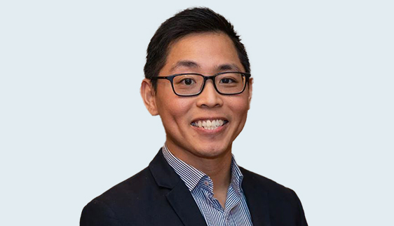 Head shot (shoulders upwards) of Dr Alexander Tan, a young man wearing black-famed glasses, a black suit jacket and collared, blue/purple shirt (with top button undone)