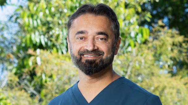 Headshot of Dr Sunil Warrier an ophthalmologist at the Vision Eye Institute Brisbane. He is wearing dark blue scrubs and is standing outside in the sun in front of local greenery and trees.