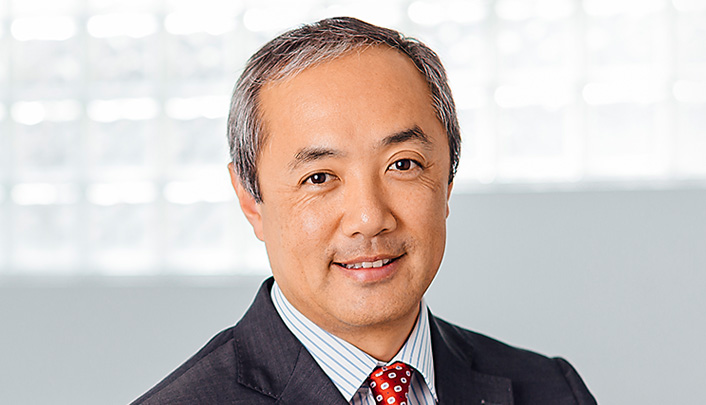 Dr David Ng dressed in a suit and tie in front of an office wall and plant