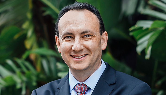 Dr Lewis Levitz dressed in a suit and tie standing in front of a wall of palm trees