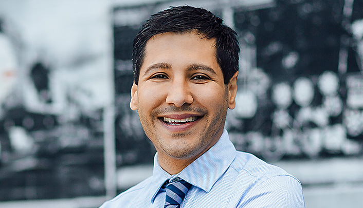 Dr Nima Pakrou dressed in a shirt and tie standing in front of a wall with a mural