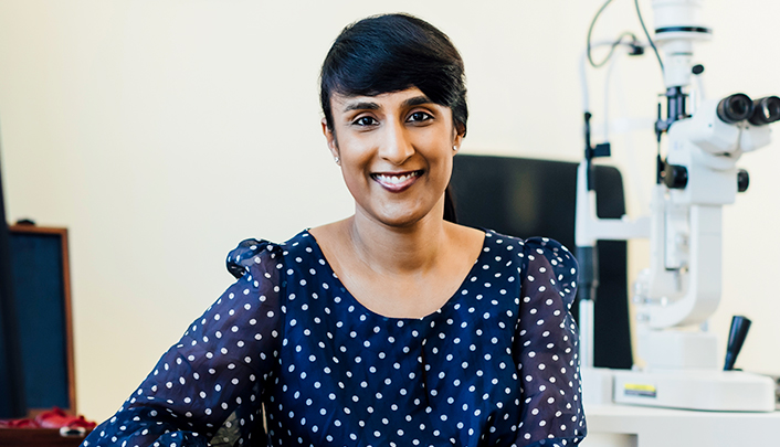 Dr Rushmia Karim (woman) in consulting suite, next to a microscope, wearing a navy blue dress with white polka dots; hair in a ponytail