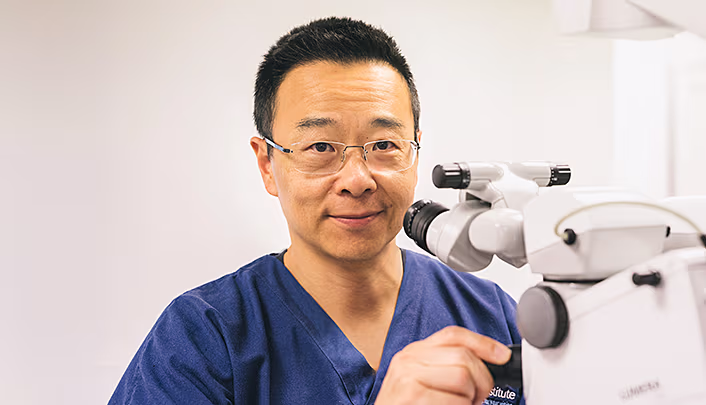 Dr Simon Chen standing in the operating room with equipment dressed in dark blue surgical scrubs and glasses