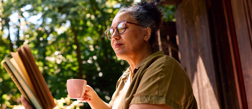 Senior woman relaxing and reading a book on a porch, enjoying her coffee and peaceful nature time outdoors.